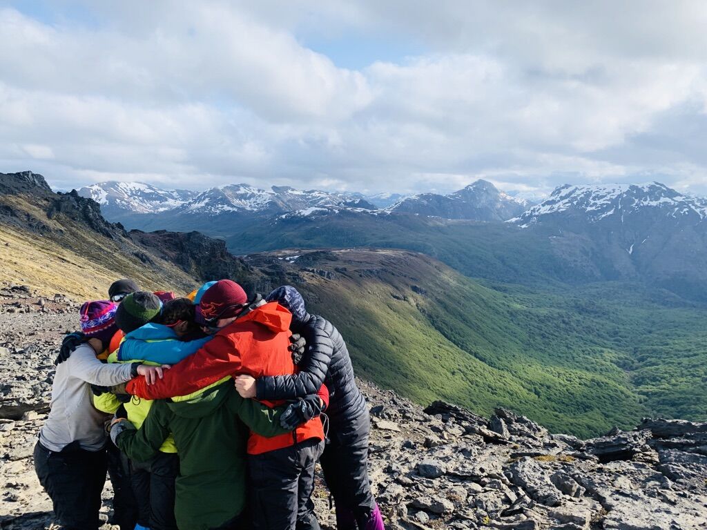 A group of people are huddled together on a rocky mountaintop, possibly celebrating a successful hike or climb. They are surrounded by a vast, scenic landscape featuring rolling green hills and snow-capped mountains in the distance under a partly cloudy sky. The overall impression is one of camaraderie and accomplishment in a beautiful natural setting.